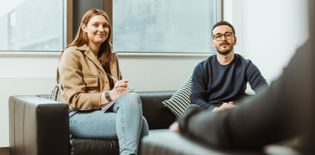 A man and a woman sat on a couch. The woman is listening to someone and making notes. 